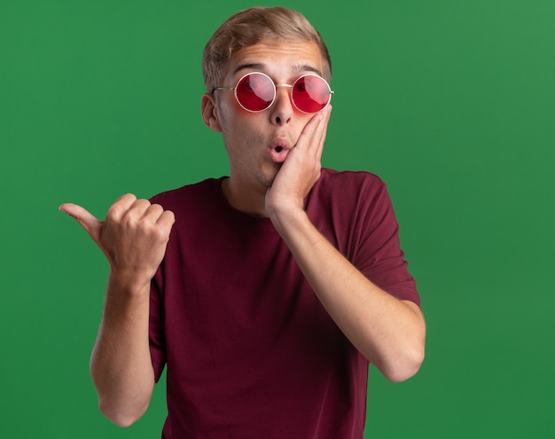 Surpris jeune beau mec portant une chemise rouge et des lunettes points sur le côté mettant la main sur la joue isolé sur le mur vert