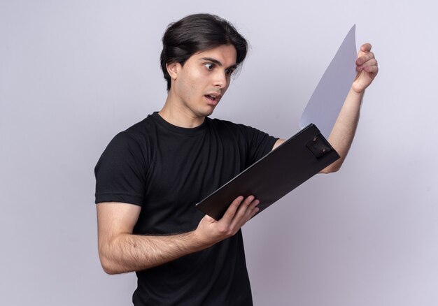 Surpris jeune beau mec portant un t-shirt noir feuilletant le presse-papiers isolé sur un mur blanc