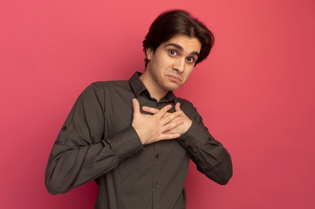 Surpris jeune beau mec portant un t-shirt noir mettant les mains sur le coeur isolé sur le mur rose