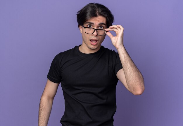 Surpris jeune beau mec portant un t-shirt noir portant et tenant des lunettes isolées sur un mur violet