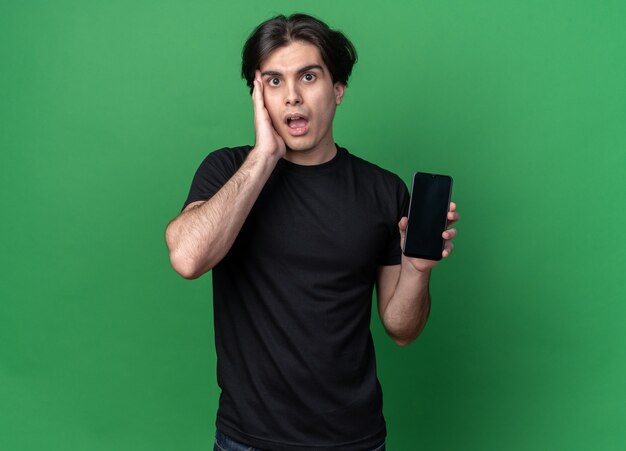 Surpris jeune beau mec portant un t-shirt noir tenant le téléphone et mettant la main sur la joue isolé sur le mur vert