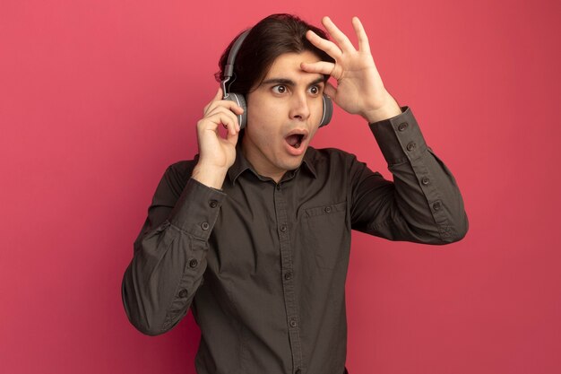 Surpris jeune beau mec vêtu d'un t-shirt noir avec des écouteurs regardant à distance avec la main isolée sur le mur rose