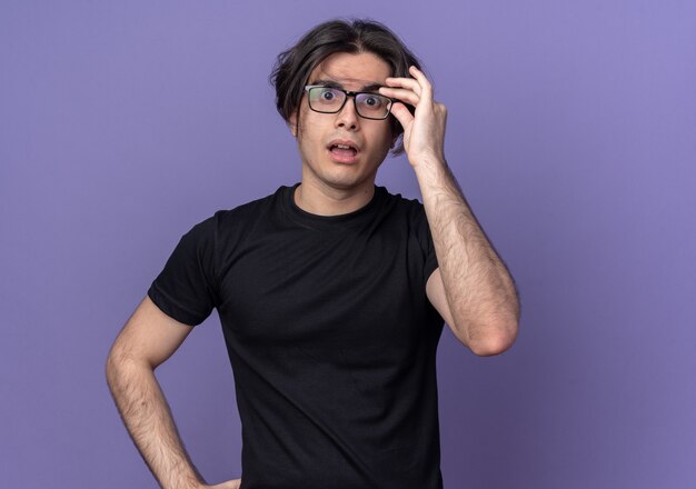 Surpris jeune beau mec vêtu d'un t-shirt noir portant et tenant des lunettes isolées sur un mur violet