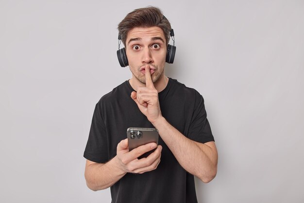 Surpris, un jeune bel homme adulte fait un geste de silence demande de se taire vêtu d'un t-shirt noir décontracté tient le téléphone portable écoute la piste audio via un casque isolé sur fond blanc.