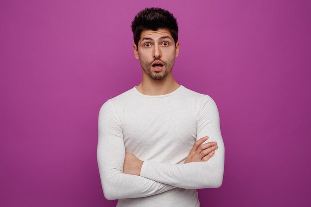 Surpris jeune bel homme regardant la caméra avec les bras croisés sur fond violet