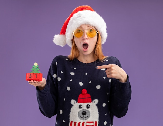 Surpris jeune belle fille portant un pull de Noël et un chapeau avec des lunettes tenant et des points au jouet de Noël isolé sur fond violet