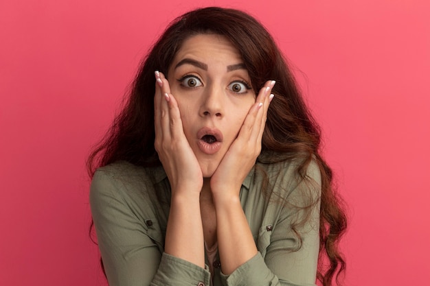 Photo gratuite surpris jeune belle fille portant un t-shirt vert olive mettant les mains sur la joue isolé sur un mur rose