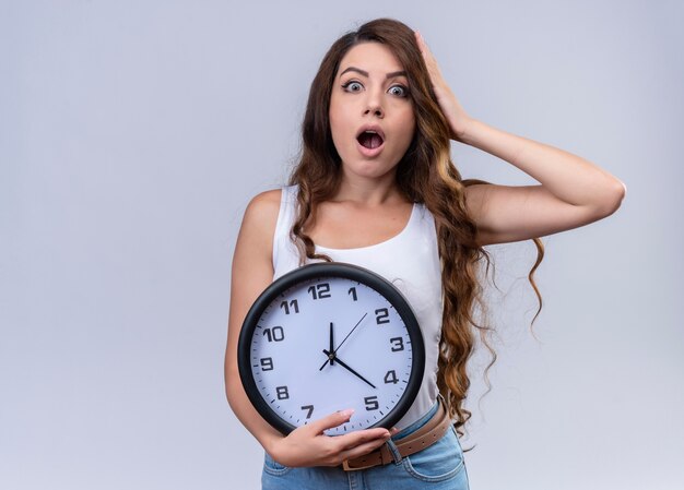 Surpris jeune belle fille tenant horloge et mettant la main sur la tête sur un mur blanc isolé avec espace copie
