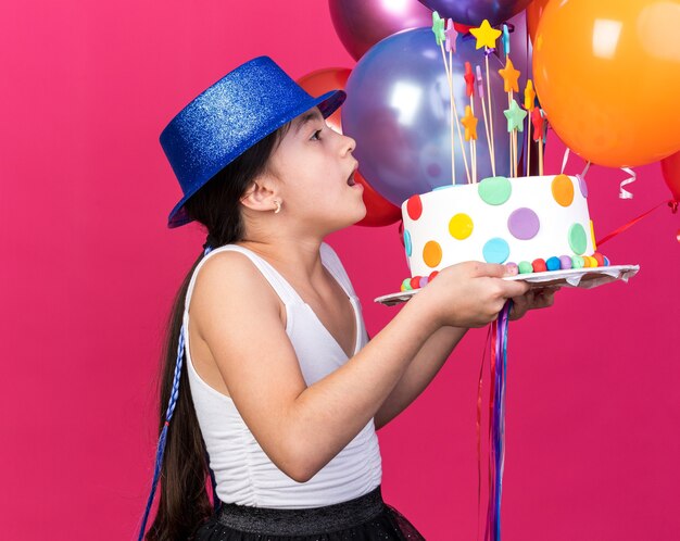 surpris, jeune, caucasien, girl, porter, bleu, chapeau partie, regarder, gâteau anniversaire, et, tenue, ballons hélium, isolé, sur, mur rose, à, espace copie