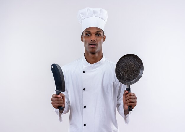 Surpris jeune cuisinier afro-américain en uniforme de chef tient le couteau et la poêle sur fond blanc isolé avec copie espace