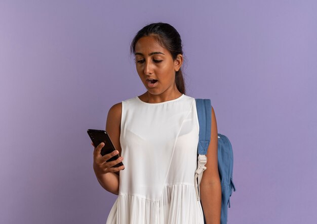 Surpris jeune écolière portant sac à dos tenant et regardant le téléphone sur violet