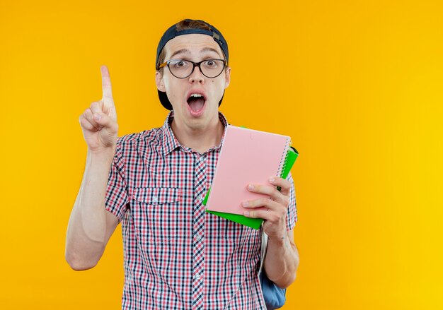 Surpris jeune étudiant garçon portant un sac à dos et des lunettes et une casquette tenant un cahier et des points sur blanc