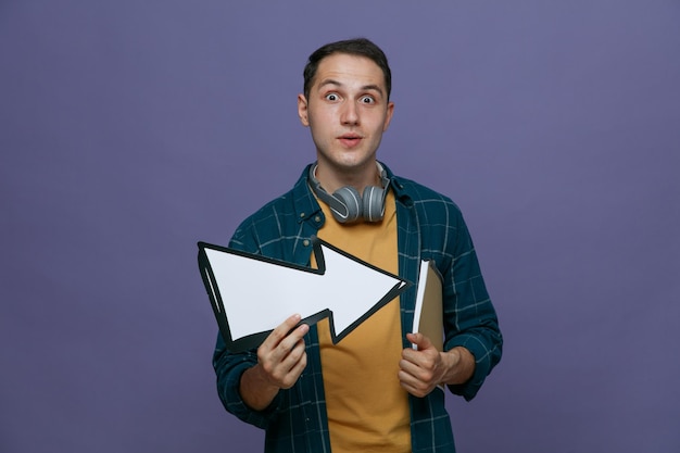 Surpris jeune étudiant masculin portant des écouteurs autour du cou tenant un bloc-notes sous le bras et une flèche pointant vers le côté regardant la caméra isolée sur fond violet