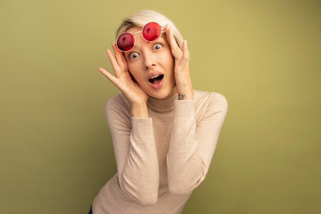 Surpris jeune femme blonde levant des lunettes de soleil regardant l'avant isolé sur un mur vert olive avec espace pour copie