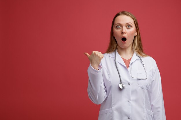 Surpris jeune femme blonde médecin portant une robe médicale et un stéthoscope autour du cou pointant vers le côté