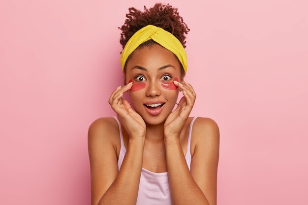 Photo gratuite surpris jeune femme bouclée à la peau sombre avec des taches de collagène sous les yeux, a l'air impressionnant, se demande un effet rajeunissant impressionnant du produit de beauté, porte un bandeau jaune et un t-shirt.