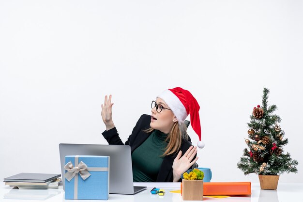 Surpris jeune femme avec chapeau de père Noël et lunettes assis à une table avec un arbre de Noël et un cadeau sur elle sur fond blanc