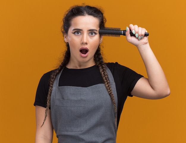 Surpris jeune femme coiffeur en uniforme mettant peigne sur temple isolé sur mur orange