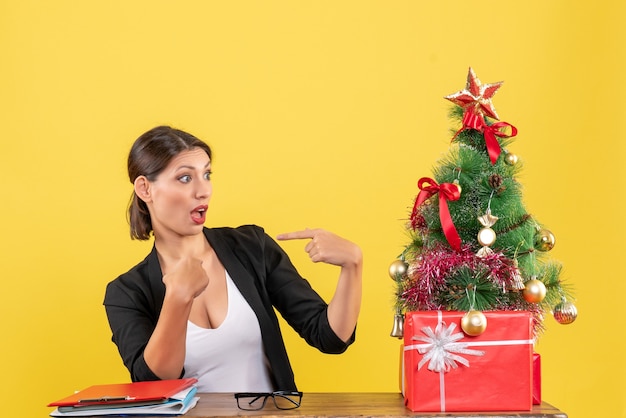 Surpris jeune femme en costume se pointant près de l'arbre de Noël décoré au bureau sur jaune