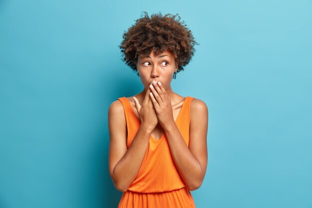 Surpris jeune femme couvre la bouche regarde impressionné entend des secrets porte une robe orange étant choqué par quelque chose pose contre le mur bleu