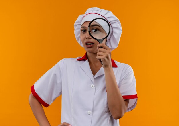 Surpris jeune femme cuisinier portant l'uniforme de chef regardant la caméra à travers la loupe sur fond orange isolé