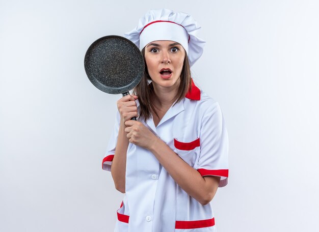 Surpris jeune femme cuisinier portant des uniformes de chef tenant une poêle à frire isolé sur fond blanc