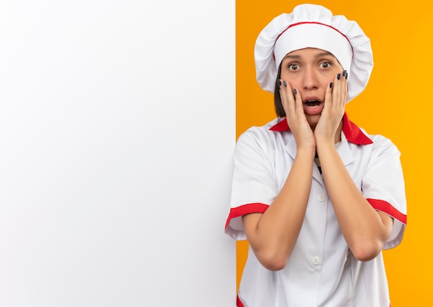 Surpris jeune femme cuisinier en uniforme de chef debout devant un mur blanc mettant les mains sur le visage isolé sur mur orange