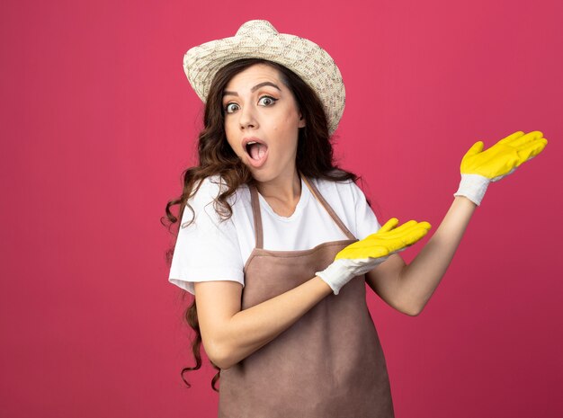 Surpris jeune femme jardinière en uniforme portant chapeau de jardinage et des gants points sur le côté avec deux mains isolé sur un mur rose avec copie espace