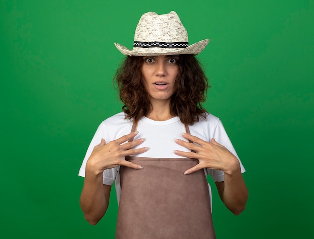 Photo gratuite surpris jeune femme jardinière en uniforme portant chapeau de jardinage mettant les mains sur la poitrine