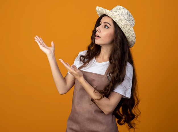 Surpris jeune femme jardinière en uniforme portant chapeau de jardinage regarde et pointe sur le côté isolé sur un mur orange avec copie espace