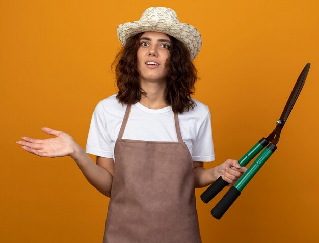 Surpris jeune femme jardinière en uniforme portant chapeau de jardinage tenant des tondeuses et répandre la main