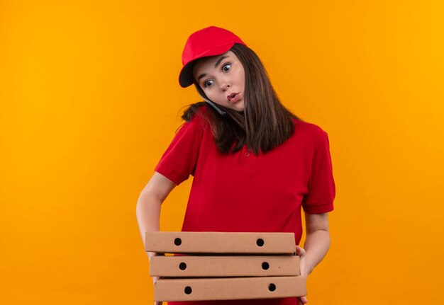 Surpris jeune femme de livraison portant un t-shirt rouge en bonnet rouge tenant une boîte à pizza et et faire un appel téléphonique sur mur orange isolé