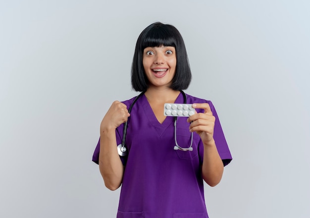 Surpris jeune femme médecin brune en uniforme avec stéthoscope garde le poing et détient la médecine