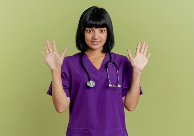 Surpris jeune femme médecin brune en uniforme avec stéthoscope se dresse avec les mains surélevées isolé sur fond vert olive avec espace copie