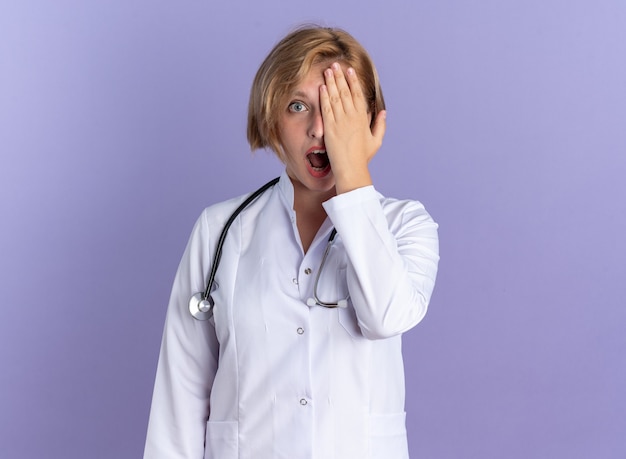 Surpris jeune femme médecin portant une robe médicale avec un œil couvert de stéthoscope avec une main isolée sur fond bleu