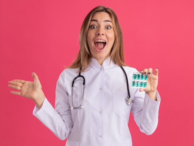 Surpris jeune femme médecin portant une robe médicale avec stéthoscope holding pills propagation main isolé sur mur rose
