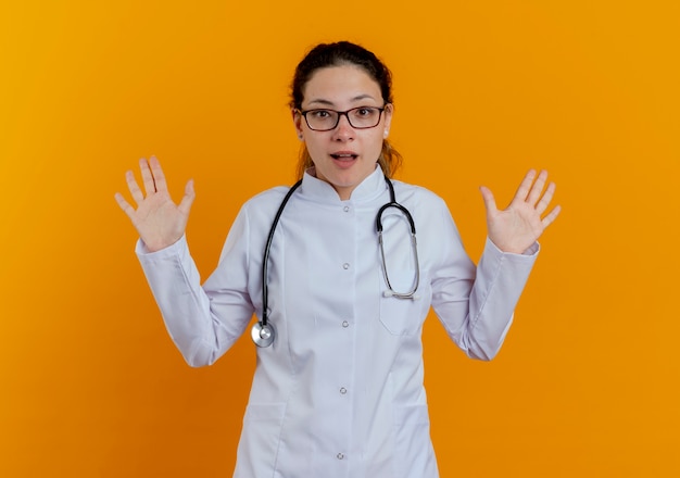 Surpris jeune femme médecin portant une robe médicale et un stéthoscope avec des lunettes écartant les mains isolées