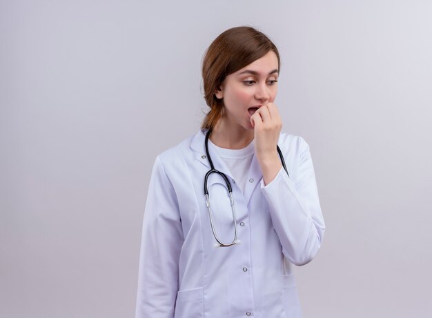 Surpris jeune femme médecin portant une robe médicale et un stéthoscope et mettant la main sur la bouche sur un mur blanc isolé avec copie espace