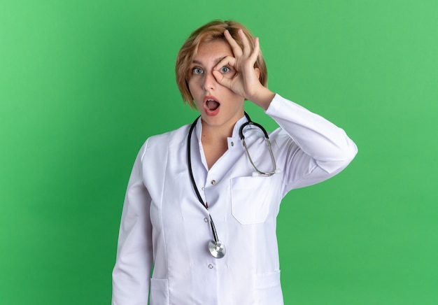 Surpris jeune femme médecin portant une robe médicale avec stéthoscope montrant un geste de regard isolé sur fond vert