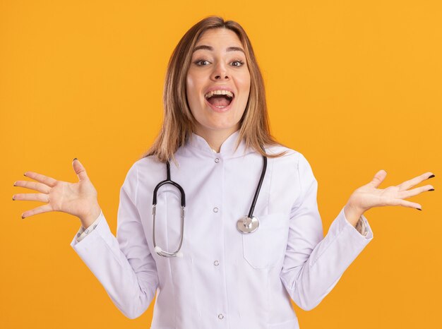 Surpris jeune femme médecin portant une robe médicale avec stéthoscope répandant les mains isolées sur le mur jaune