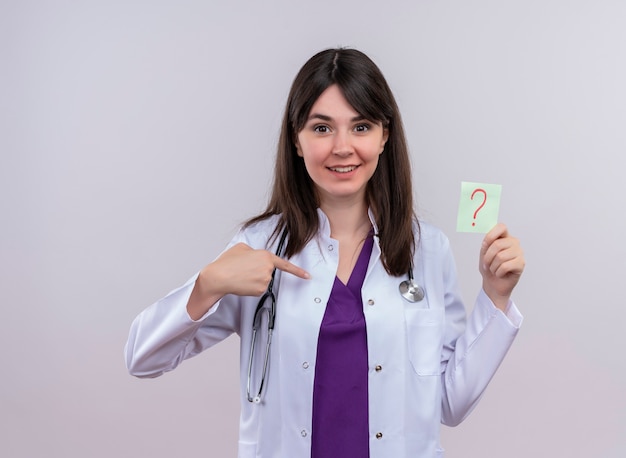 Surpris jeune femme médecin en robe médicale avec stéthoscope pointe sur elle-même et détient un point d'interrogation sur fond blanc isolé avec copie espace