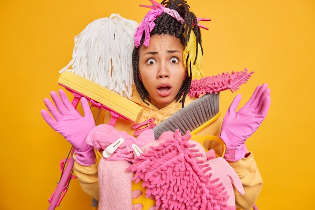 Surpris, une jeune femme à la peau foncée avec des dreadlocks pose près d'un seau plein de trucs de nettoyage de maison