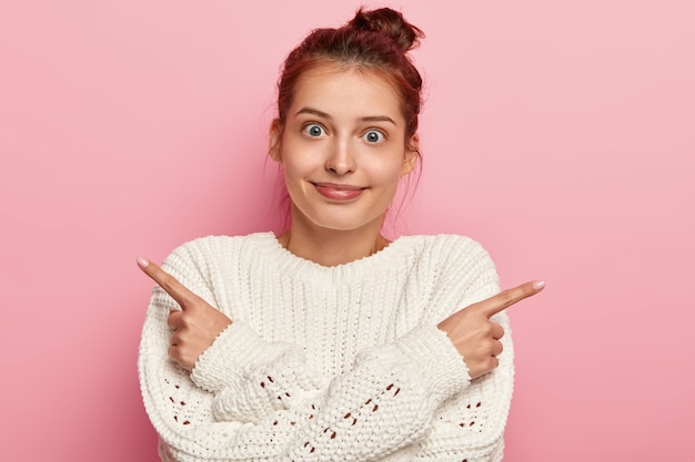 Surpris, une jeune femme pointe sur le côté, choisit entre deux options, porte un pull d'hiver blanc, a les cheveux peignés, pose sur fond rose, ne peut pas faire de choix, décide quoi acheter