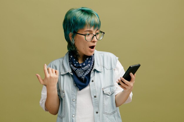 Surpris jeune femme portant des lunettes bandana sur le cou en gardant la main dans l'air à l'aide de téléphone portable isolé sur fond vert olive