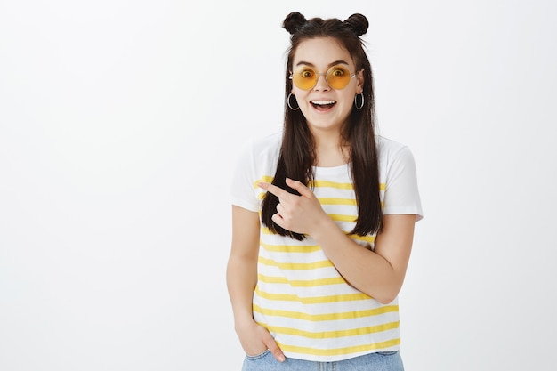 Surpris jeune femme posant avec des lunettes de soleil contre le mur blanc