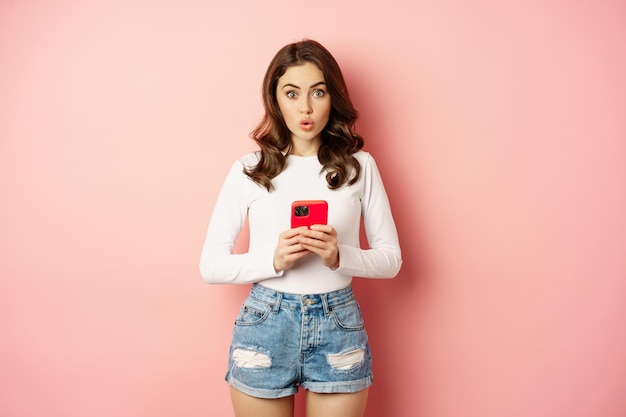 Surpris jeune femme tenant un smartphone, regardant la caméra étonnée, vérifiant la publicité, l'annonce d'achat en ligne, debout sur fond rose