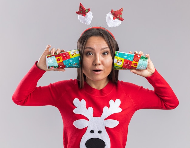 Photo gratuite surpris jeune fille asiatique portant cerceau de cheveux de noël avec chandail mettant la tasse de noël sur les oreilles isolé sur fond blanc