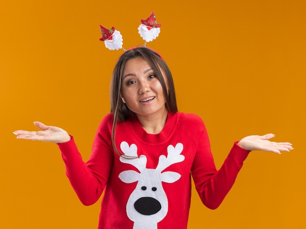 Surpris jeune fille asiatique portant cerceau de cheveux de Noël avec chandail répandant les mains isolé sur mur orange