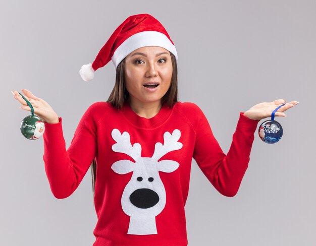 Surpris jeune fille asiatique portant chapeau de Noël avec chandail tenant des boules de sapin de Noël isolé sur fond blanc