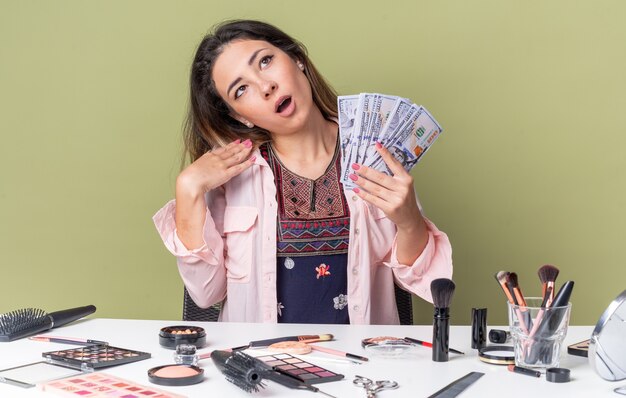 Surpris jeune fille brune assise à table avec des outils de maquillage tenant de l'argent et levant les yeux